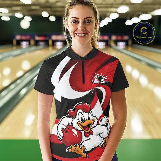 A woman standing in a bowling alley wearing a custom short-sleeve bowling shirt with a red, black, and white graphic design featuring a cartoon rooster holding a bowling ball. The shirt has a black collar and a short black zipper at the neck.
