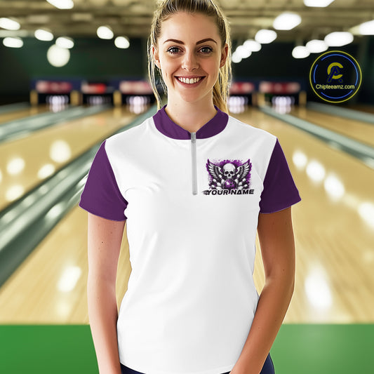 A woman stands in a bowling alley wearing a bowling shirt with quarter zip collar, purple skull-themed bowling graphic with black wings, pins, bowling ball