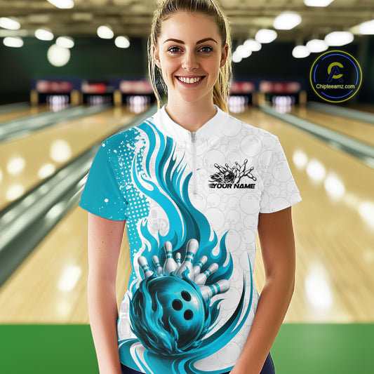 A woman stands in a bowling alley wearing a short-sleeve bowling shirt with a white base, teal and blue flame-like swirls, a large bowling ball graphic near the hem, and a black chest logo that reads 'YOUR NAME'.