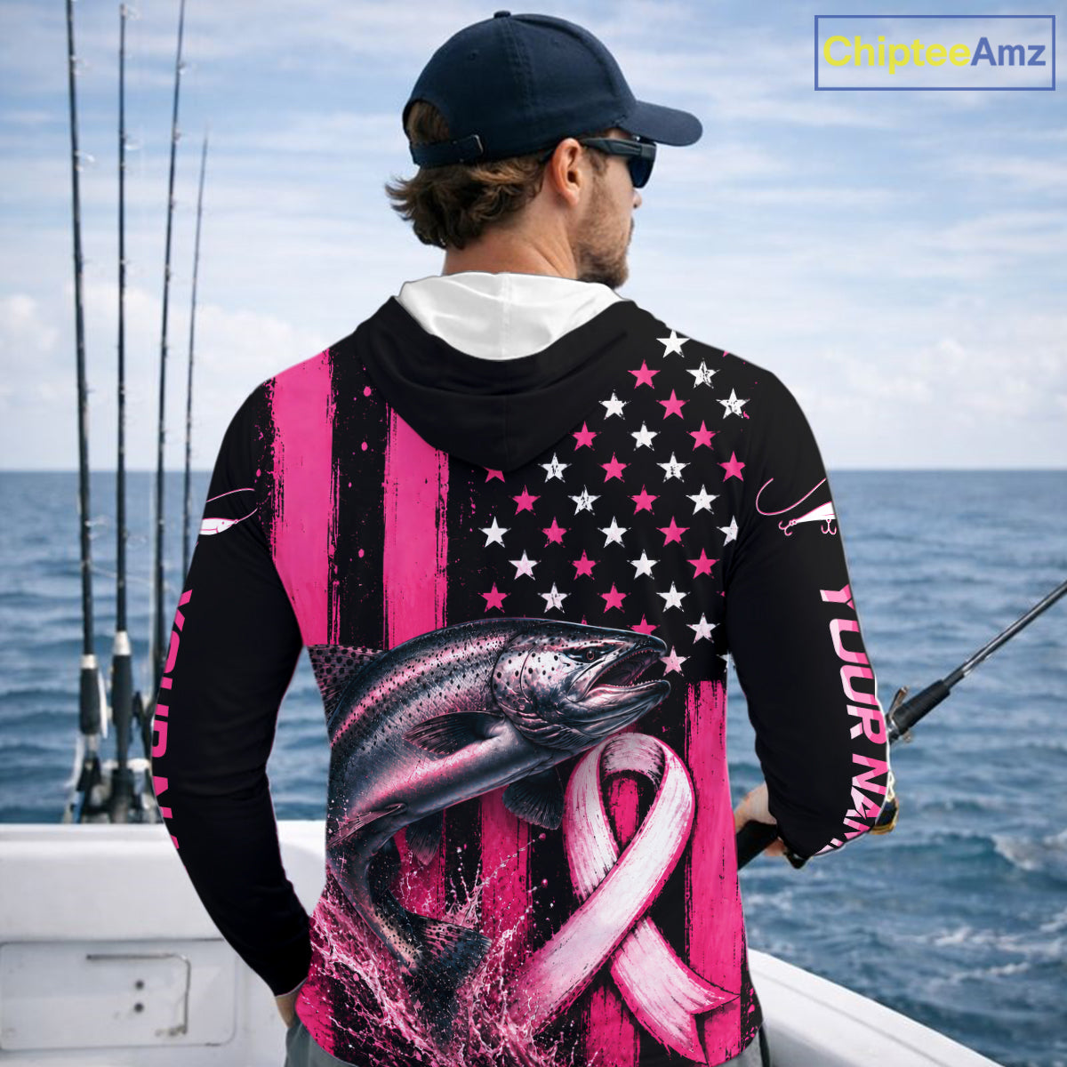 A man on a boat wearing a black long-sleeve hoodie with a pink and white patriotic fish graphic on the back, featuring stars, stripes, and a pink ribbon. He is wearing a dark blue cap and sunglasses, with fishing rods visible in the background.