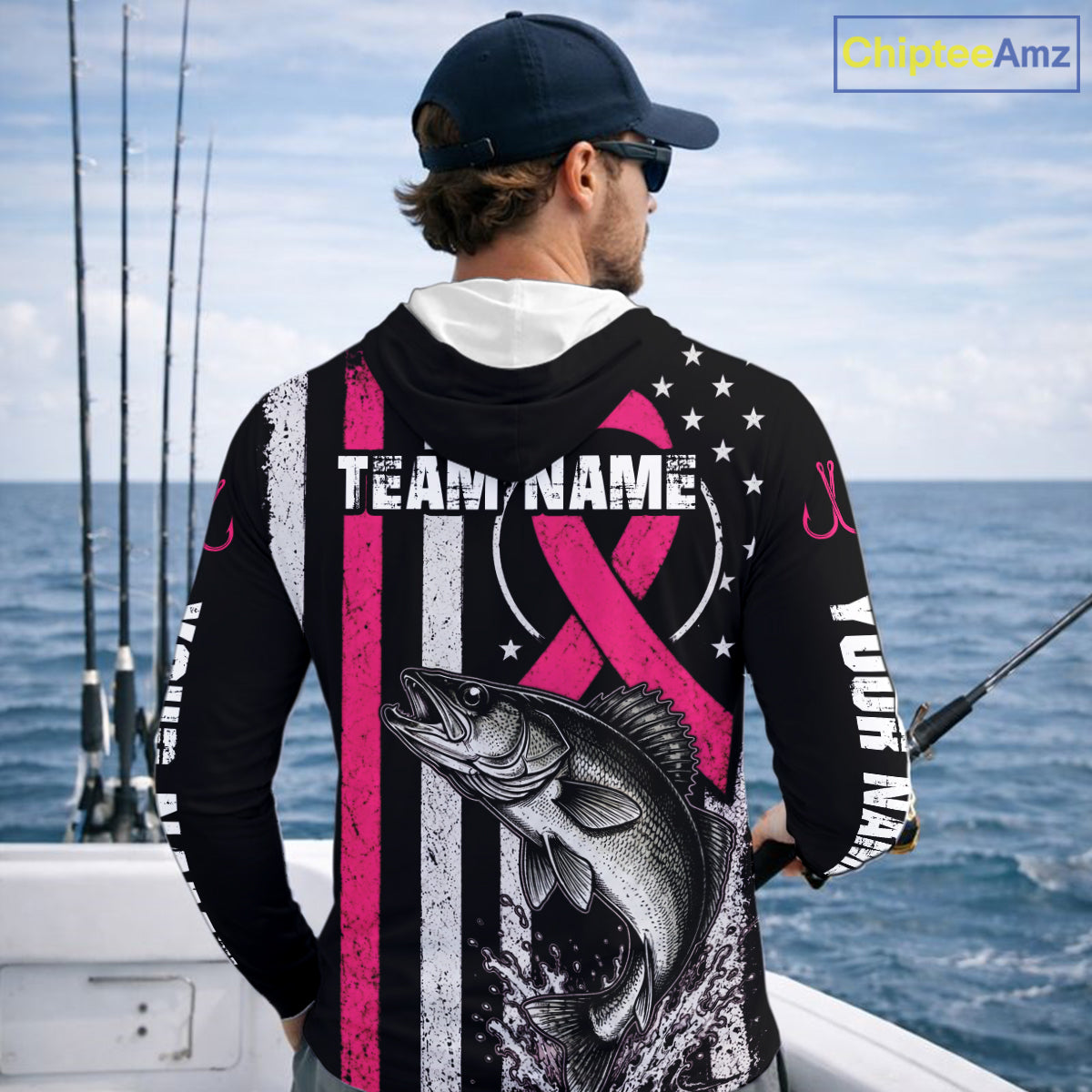 A man on a boat shown from behind wearing a custom black long-sleeve fishing hoodie with a pink and white striped design, a large bass graphic, and customizable text. The background shows the ocean and fishing rods.