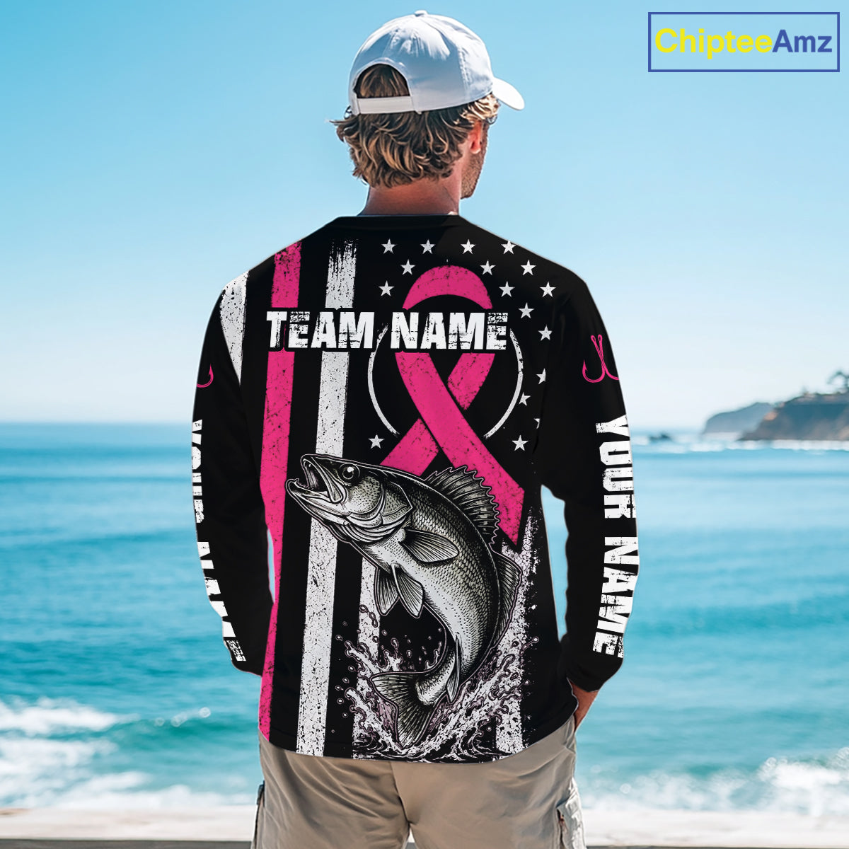 A man standing on a beach viewed from behind, wearing a black long-sleeve shirt with a large pink and white graphic design featuring a fish, stars, stripes, and customizable text. The background shows the blue ocean and sky.