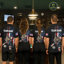 Load image into Gallery viewer, A back view of a team of four pool players standing in a line in front of a blue felt pool table in a billiards room. All four are wearing the back view of the custom black billiards polo shirt, which features a large distressed US flag pattern, a large white 'TEAM NAME' text placeholder, and a cat and 8-ball motif. 