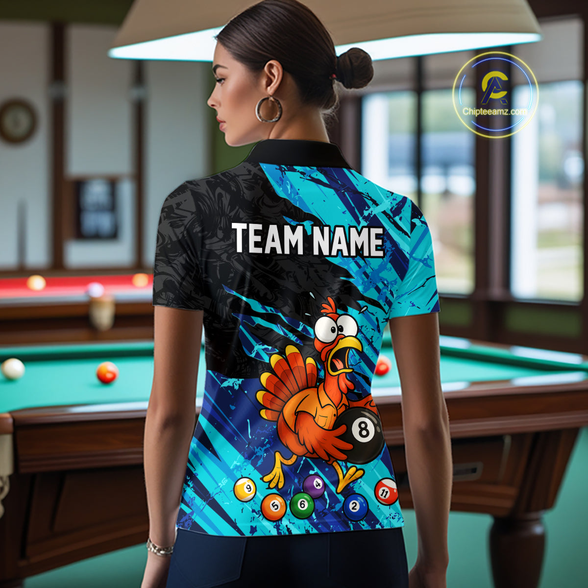 A woman stands in a billiards room wearing a custom-printed short-sleeve polo shirt. The back of the shirt features a black and cyan abstract background with the text 'TEAM NAME' and a cartoon turkey holding an 8-ball pool cue.