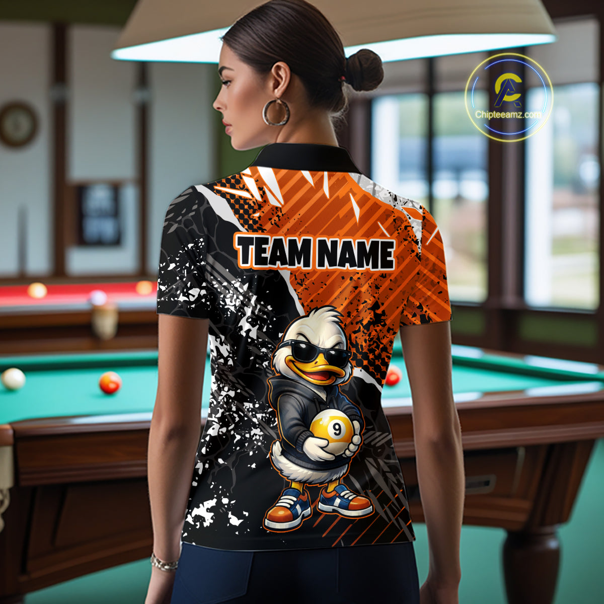 A woman stands in a billiards room wearing a custom-printed short-sleeve polo shirt. The shirt features a split design with a black and white splatter pattern on one side and a bright orange patterned side, with a cartoon duck mascot holding a billiard ball and the text 'TEAM NAME' printed on the back.