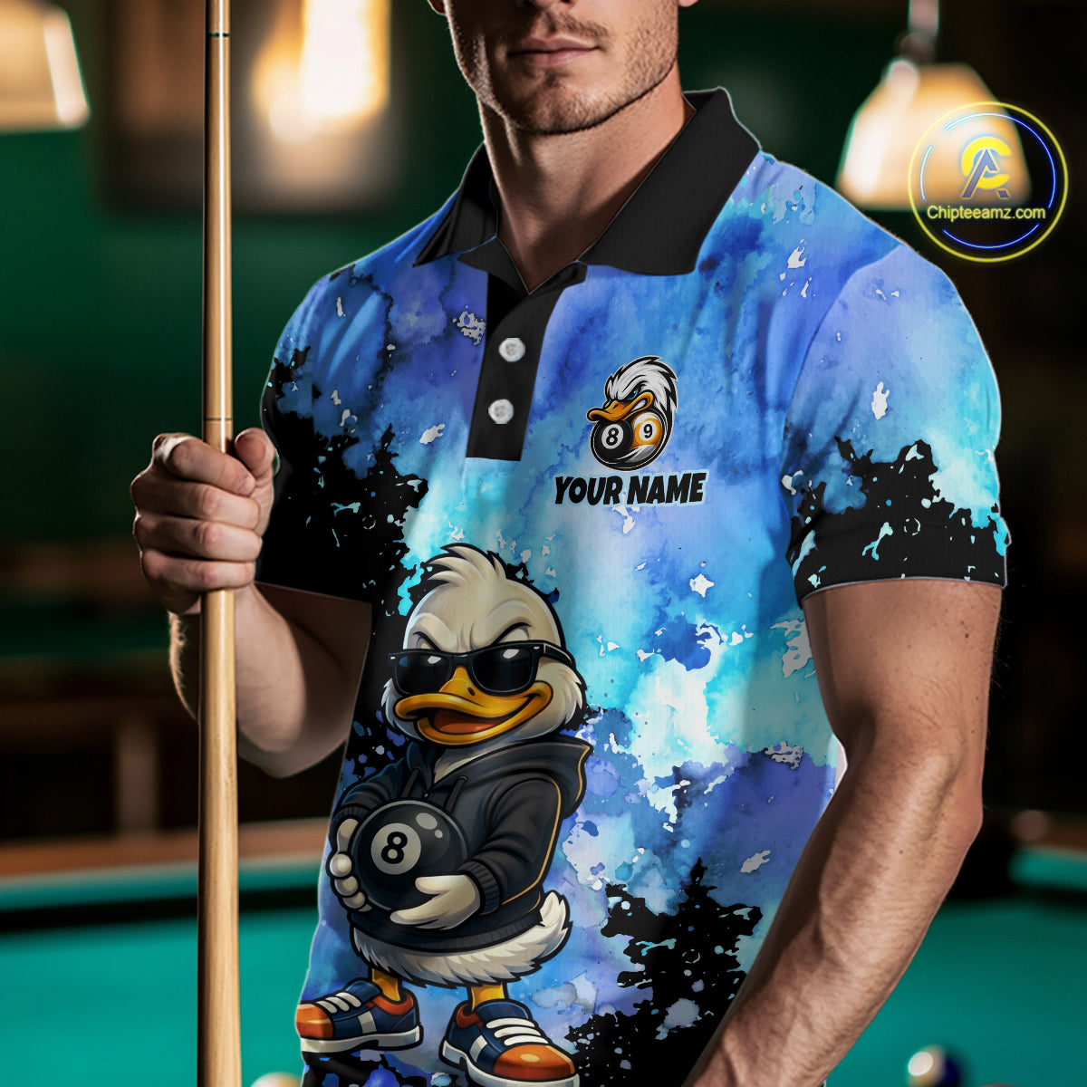A man wearing a custom blue and black watercolor-patterned polo shirt with a cartoon duck graphic holding an 8-ball and the text 'YOUR NAME'. He is holding a pool cue in a dimly lit pool hall environment.