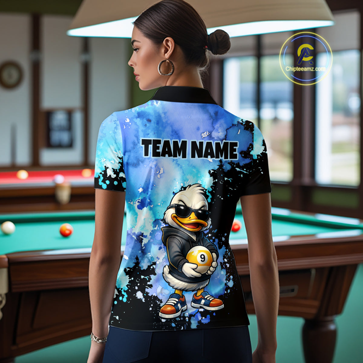 A woman stands in a billiards room wearing a custom-printed short-sleeve polo shirt from the back. The shirt features a blue and black watercolor splatter design with a cartoon duck mascot holding a yellow billiard ball and the text 'TEAM NAME'.