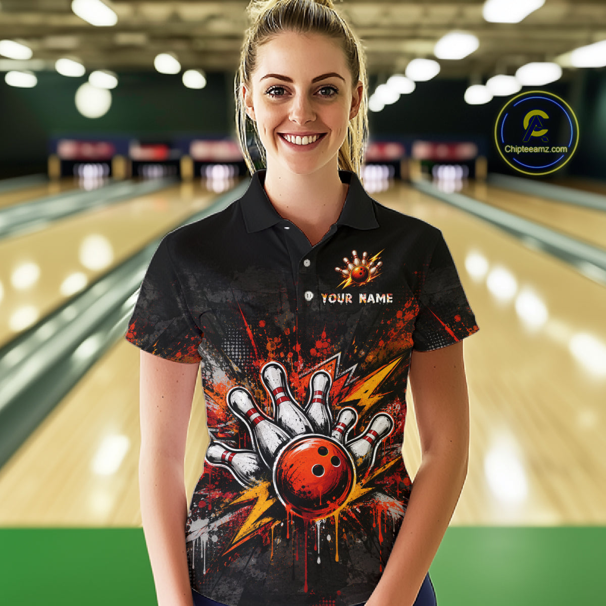 A woman stands in a bowling alley wearing a black short-sleeve polo shirt with a colorful bowling-themed graphic on the front. The shirt features a red bowling ball, white bowling pins, orange splatter effects, and the text 'YOUR NAME' on the chest.