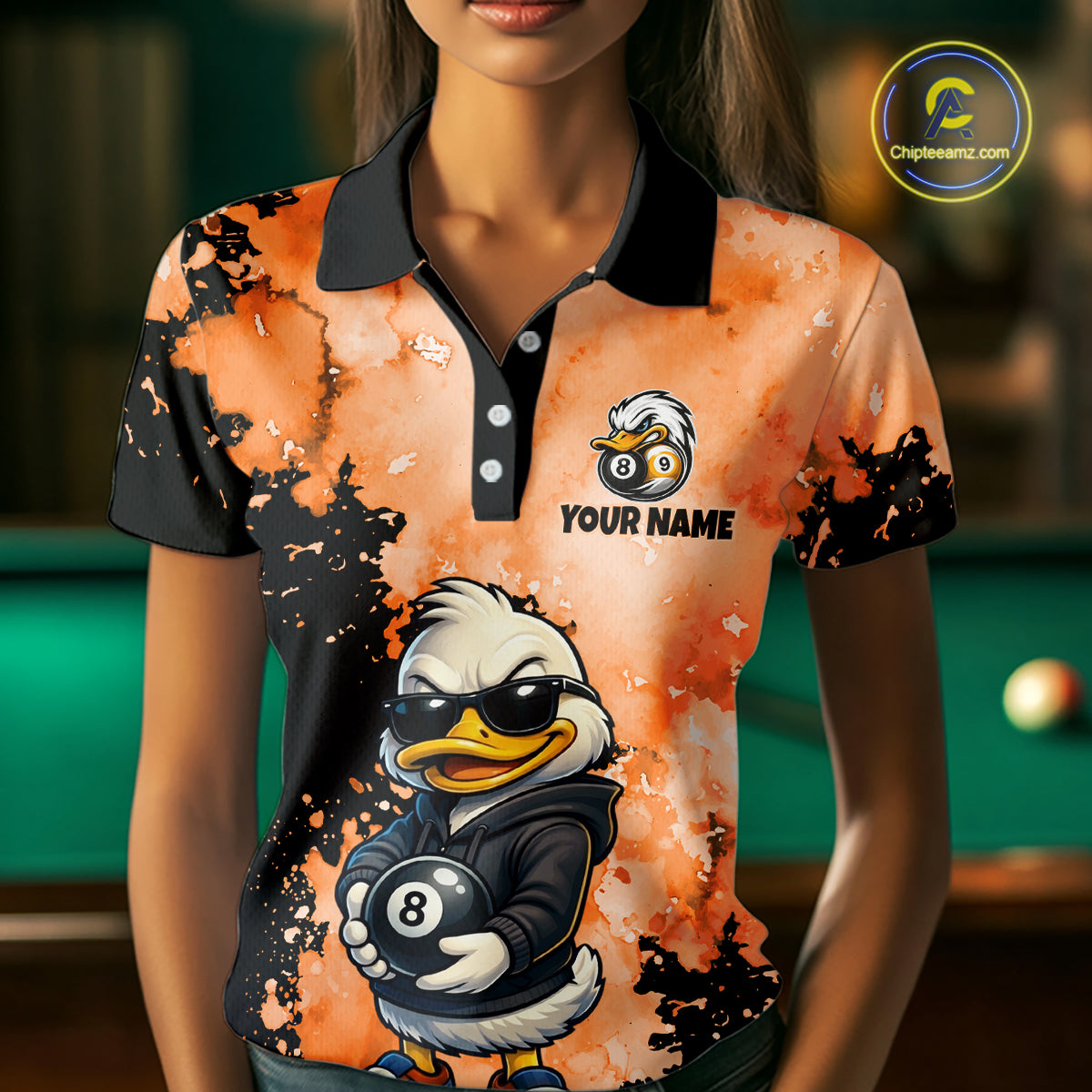 A woman wearing a custom-printed polo shirt with a black collar and sleeves, featuring an orange and black splatter design, a cartoon duck wearing sunglasses and a black jacket holding an 8-ball, and the text 'YOUR NAME'.