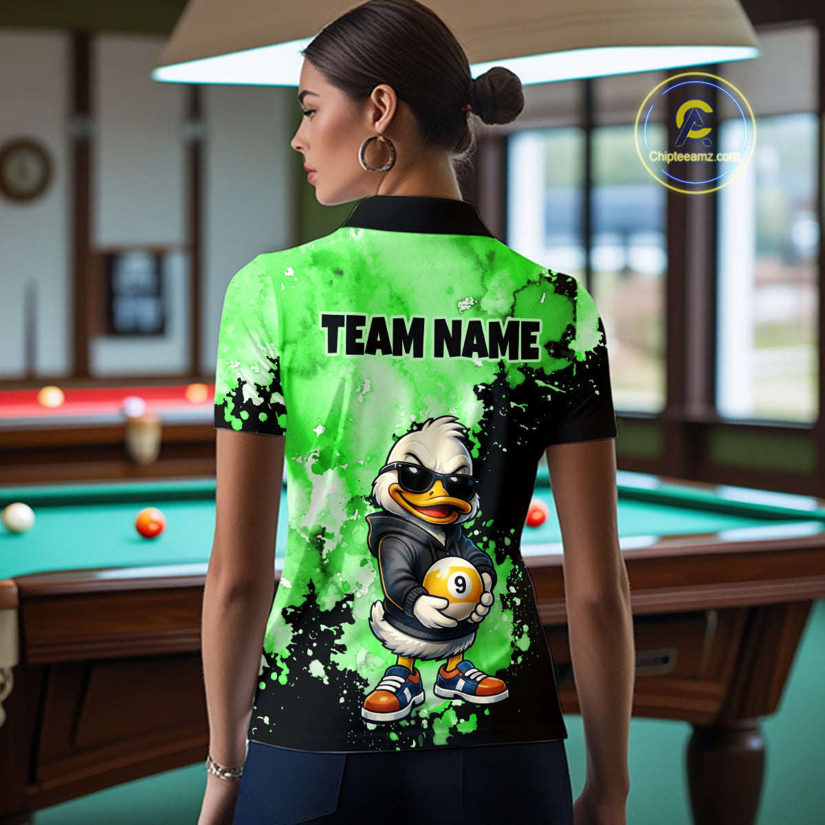 A woman stands in a billiards room wearing a custom short-sleeve polo shirt from the back. The shirt features a vibrant green and black splatter design with a cartoon duck mascot holding a pool ball and the text 'TEAM NAME'.