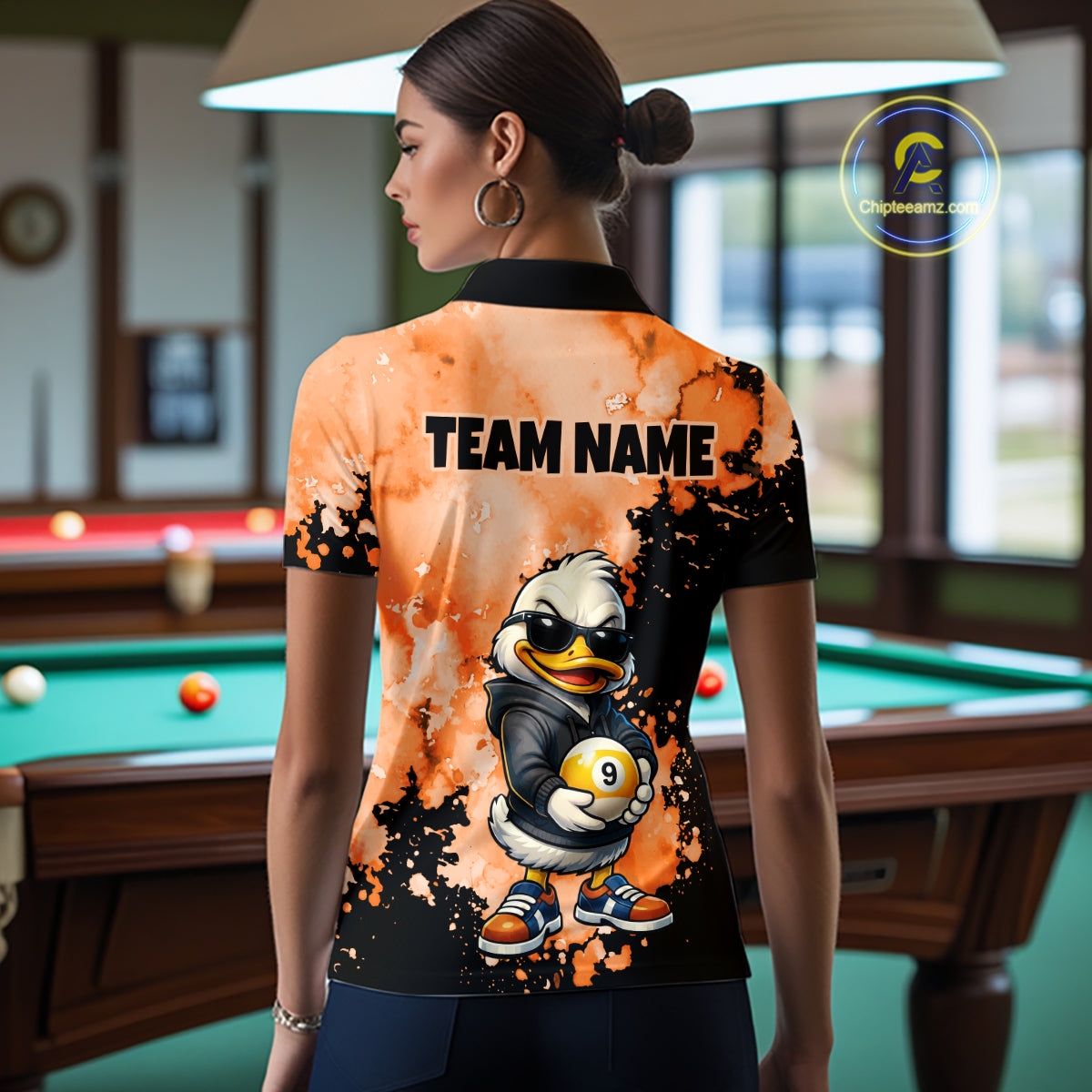 A woman stands in a billiards room wearing a custom-printed short-sleeve polo shirt from the back. The shirt features a black and orange splatter design with a cartoon duck mascot holding a billiard ball and the text 'TEAM NAME'.