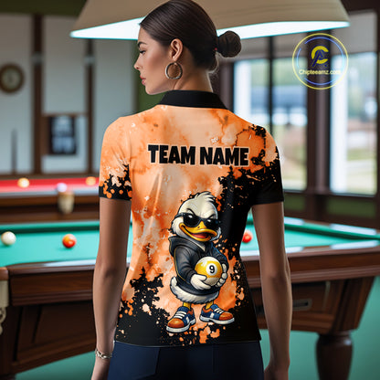 A woman stands in a billiards room wearing a custom-printed short-sleeve polo shirt from the back. The shirt features a black and orange splatter design with a cartoon duck mascot holding a billiard ball and the text 'TEAM NAME'.