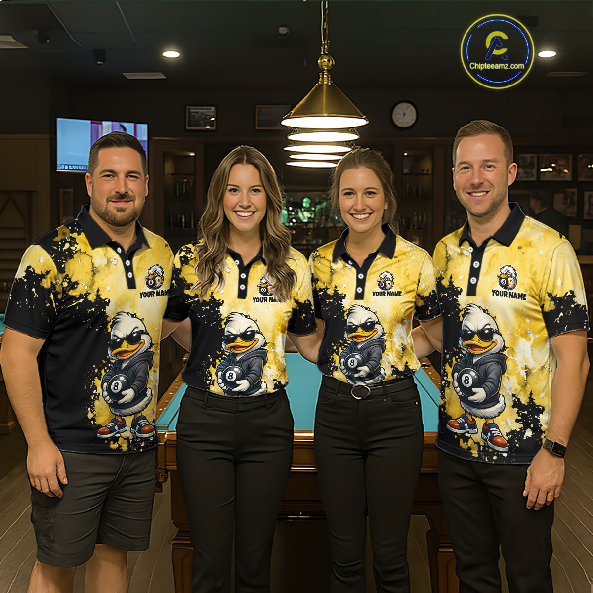 Four people standing together in a dimly lit indoor venue, wearing matching yellow and black polo shirts with a cartoon duck graphic and the text 'YOUR NAME'