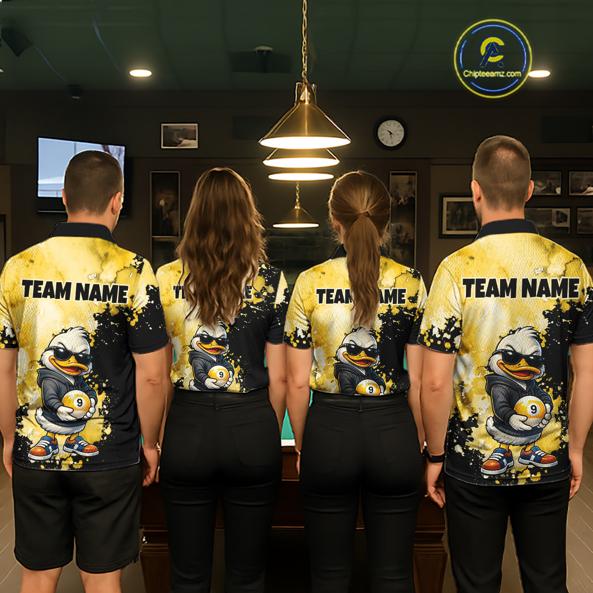 Four people standing with their backs to the camera in a dimly lit indoor room, wearing matching yellow and black custom team polo shirts with a cartoon duck graphic and 'TEAM NAME' printed across the back.