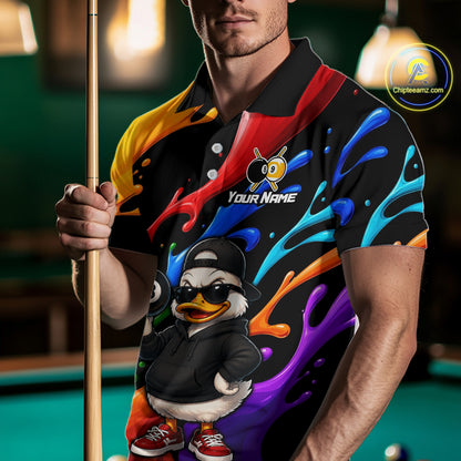 A man in a poolroom wearing a black short-sleeve polo shirt with a colorful abstract paint-splatter design and a cartoon duck character graphic on the front. He is holding a pool cue, and the shirt features a customizable 'Your Name' placeholder.