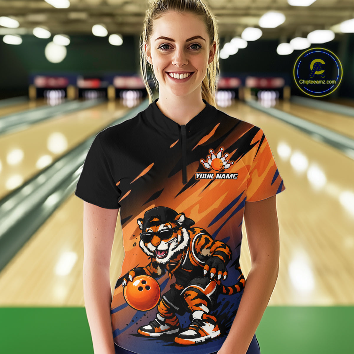 A woman stands in a bowling alley wearing a custom short-sleeve polo shirt with a black collar and a colorful front graphic featuring a tiger mascot holding an orange bowling ball. The shirt has orange, blue, and black graphics with 'YOUR NAME' printed on the chest.