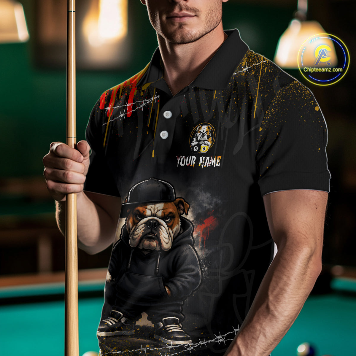 A man wearing a black custom polo shirt with a graphic of an English Bulldog in a hoodie and cap, holding a pool cue in a dimly lit pool hall. The shirt features a bulldog graphic, a custom name placeholder, and paint splatter details.