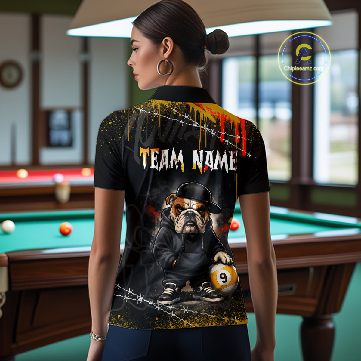 A woman stands in a billiards room wearing a black short-sleeve polo shirt with a custom graphic print. The back of the shirt features a cartoon bulldog in a black hat and jacket holding a 9-ball, with 'TEAM NAME' text and paint splatter details.