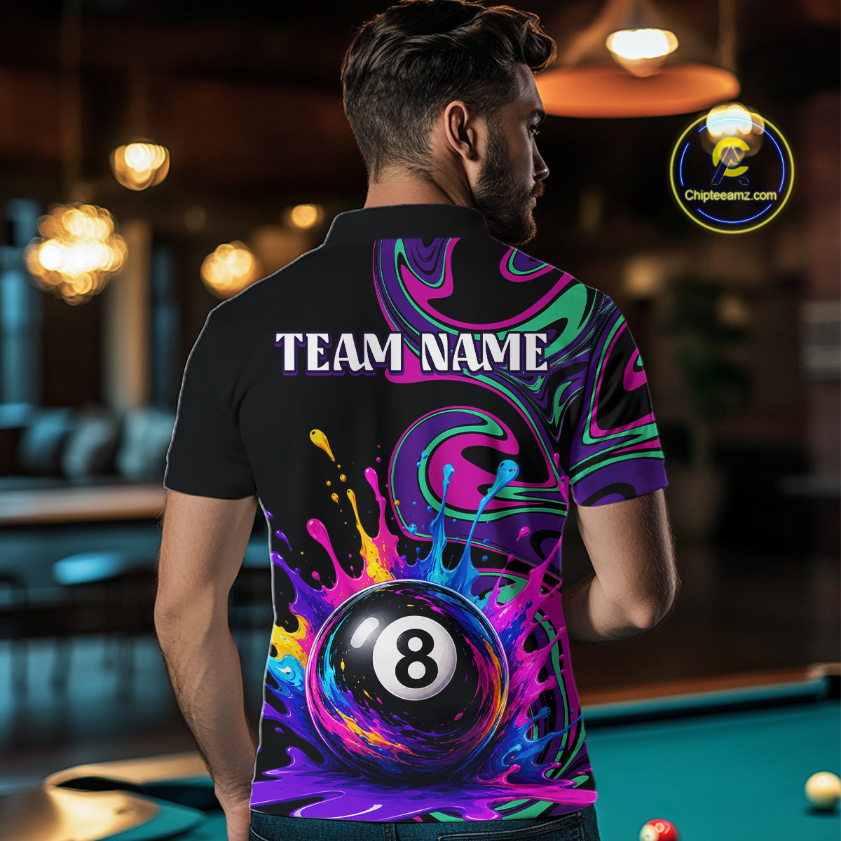 A man viewed from behind wearing a black short-sleeve polo shirt with a vibrant, colorful graphic print featuring a 8-ball pool cue surrounded by neon paint splatters and the text 'TEAM NAME'. The background is a dimly lit pool hall with a green pool table.