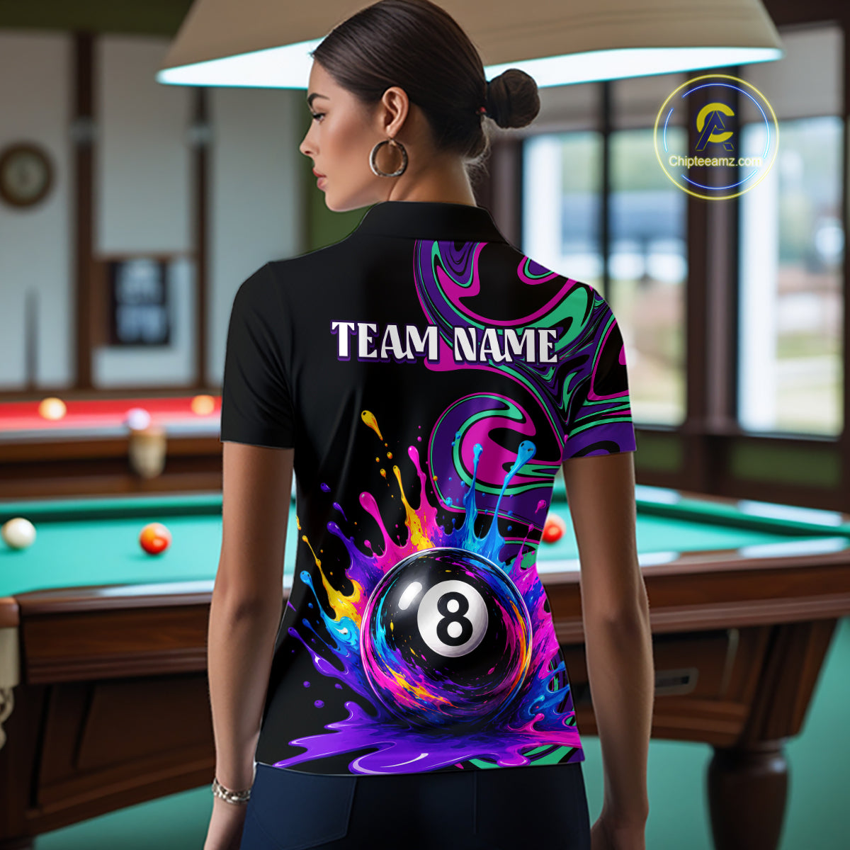 A woman stands with her back to the camera in a billiards room, wearing a black short-sleeve polo shirt with a vibrant, colorful graphic print featuring a 8-ball and the text 'TEAM NAME'.