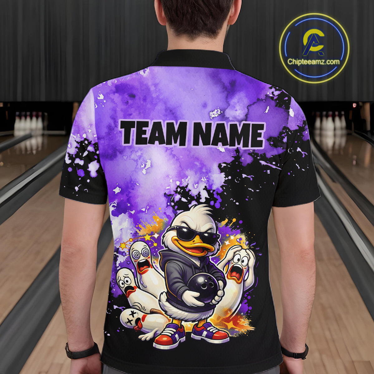 A person viewed from behind wearing a black short-sleeve bowling-themed polo shirt with a large purple and black graphic print featuring cartoon ducks, flames, and the text 'TEAM NAME'. The background shows bowling lanes.