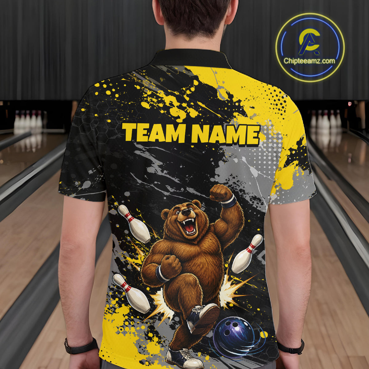 A person standing in a bowling alley viewed from behind, wearing a short-sleeve custom bowling shirt with a bold graphic design. The shirt features a brown bear bowling, white pins, a blue bowling ball, and yellow 'TEAM NAME' text on a black and yellow splatter background.