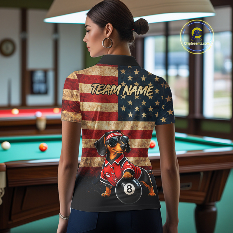 A woman stands in a billiards room wearing a custom short-sleeve polo shirt from the back. The shirt features a distressed American flag design with gold stars, a cartoon dachshund wearing a red racing jacket and goggles holding a billiard 8-ball, and the text 'TEAM NAME'.