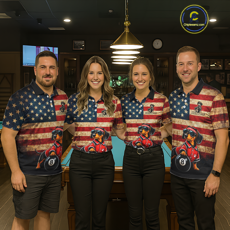 Four people standing together in a dimly lit indoor venue, wearing matching polo shirts with a distressed American flag design and a graphic of a dachshund wearing sunglasses and a red shirt, holding a pool table.