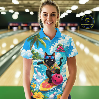 A woman stands in a bowling alley wearing a bright blue short-sleeve polo shirt with a colorful graphic print of a black cat surfing on a yellow board with a pink bowling ball, set against a blurred bowling lane background.