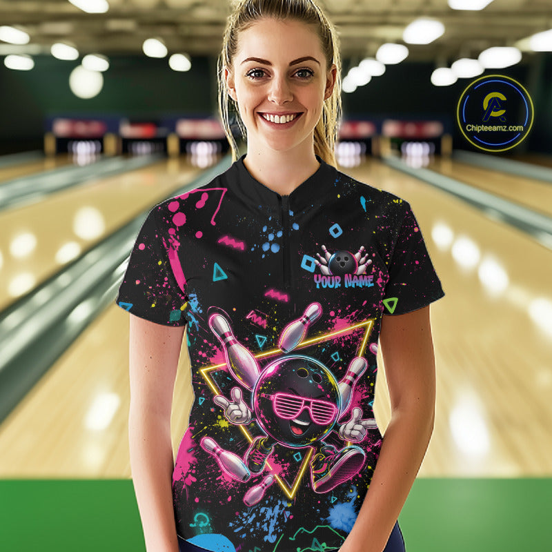 A woman standing in a bowling alley wearing a black short-sleeve bowling shirt with a colorful neon bowling graphic on the front. The shirt features a bowling pin and bowling ball design in pink, blue, and yellow, with a 'YOUR NAME' placeholder on the chest.
