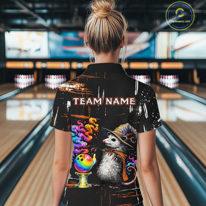 A woman viewed from behind wearing a black short-sleeve bowling-themed t-shirt with a colorful graphic of a opossum in a wizard hat next to a bowling ball and the text 'TEAM NAME'. The background shows blurred bowling alley lanes and screens.