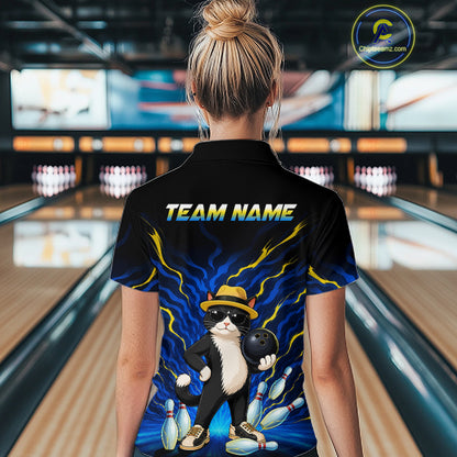 A woman viewed from behind wearing a black short-sleeve bowling-themed polo shirt with a cartoon cat in a hat holding a bowling ball, blue lightning graphics, and the text 'TEAM NAME' on the back. The background shows a blurred bowling alley interior.