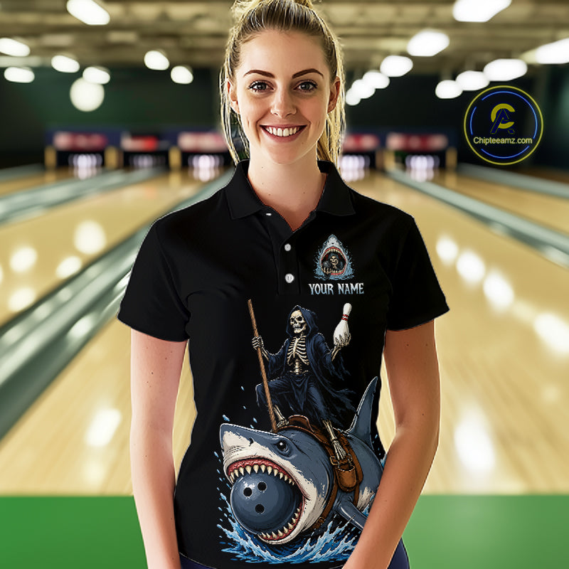 A woman stands in a bowling alley wearing a black short-sleeve polo shirt with a large graphic print of a skeleton riding a shark holding bowling pins. The shirt has a customizable design area on the chest with the text 'YOUR NAME' and an icon.