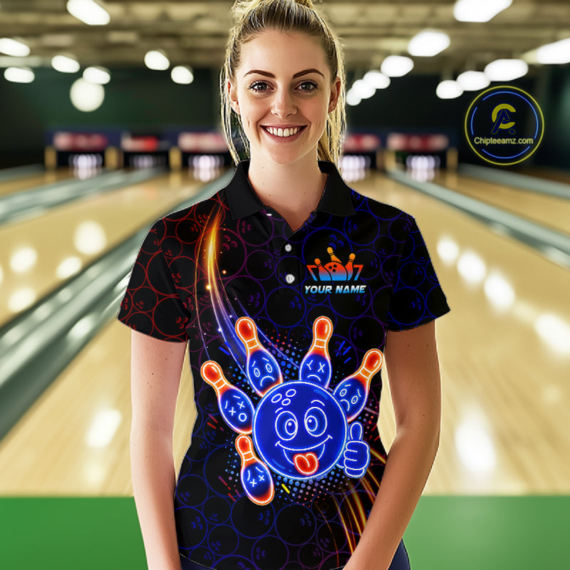 Woman standing in a bowling alley wearing a black neon-style bowling polo shirt with a glowing blue cartoon bowling ball mascot, fiery orange pin graphics, and custom name text.