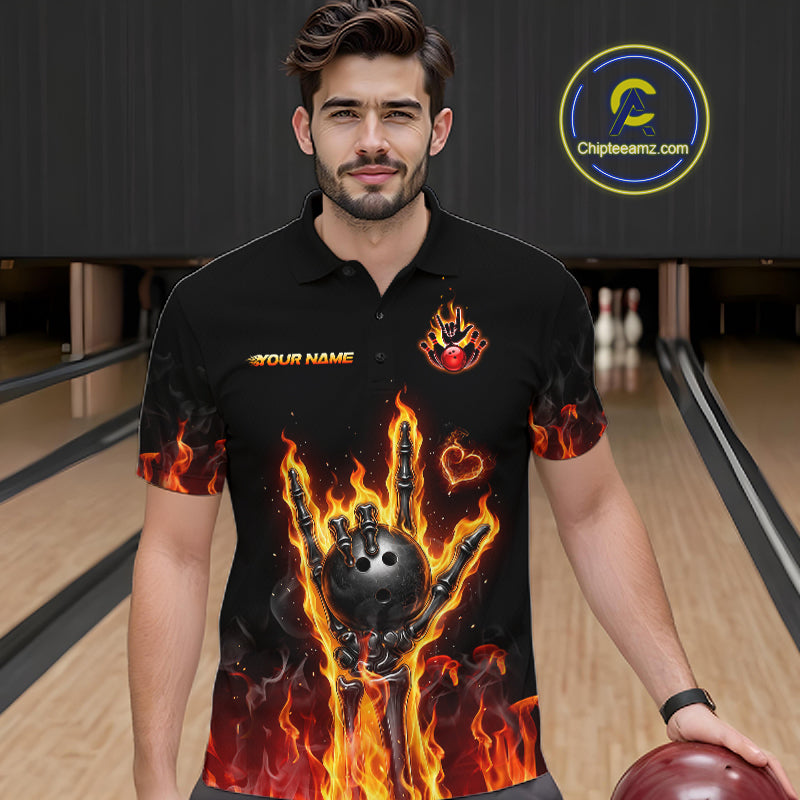 A man wearing a black short-sleeve polo shirt with a fiery flame-bomb bowling graphic, standing in a bowling alley. The shirt features orange and yellow flames, a black bowling ball and claw hand, and the text 'YOUR NAME' on the chest.
