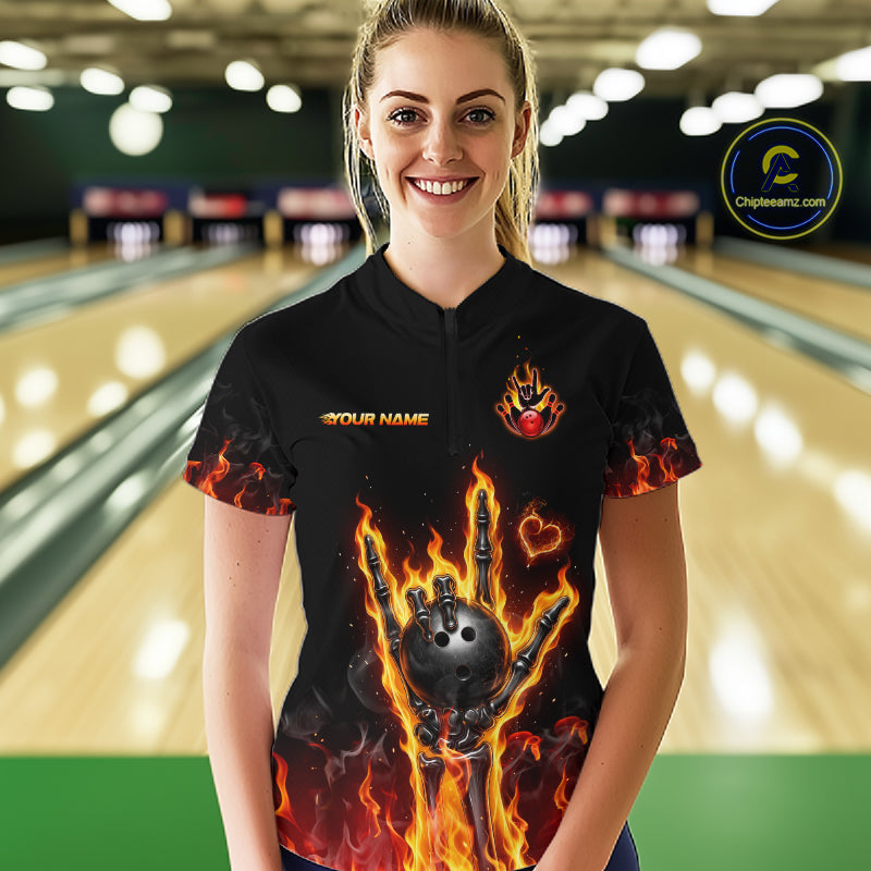 A woman stands in a bowling alley wearing a black short-sleeve bowling shirt with a fiery graphic of a bowling ball and flames, along with 'YOUR NAME' and a logo on the chest. 
