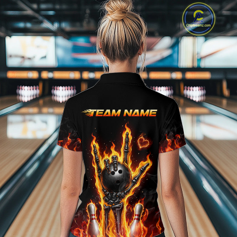 A woman viewed from behind wearing a black short-sleeve bowling shirt with a large graphic print of a flaming bowling ball and pins, with 'TEAM NAME' printed across the upper back. The background shows a blurred bowling alley.