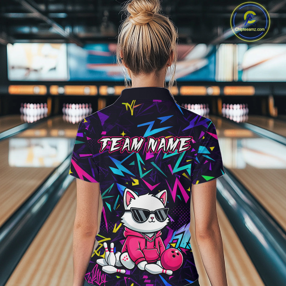 Woman wearing a black bowling polo with neon geometric patterns, custom team text, and a white cat mascot in sunglasses holding a pink bowling ball.