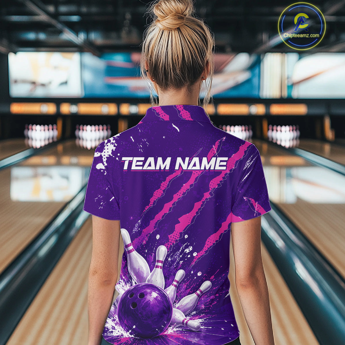 A woman viewed from behind wearing a purple bowling-themed short-sleeve polo shirt with pink claw-stripe graphics, a bowling ball and pins print on the back, and the text 'TEAM NAME' across the upper back. The background shows a blurred bowling alley with lanes and pins.