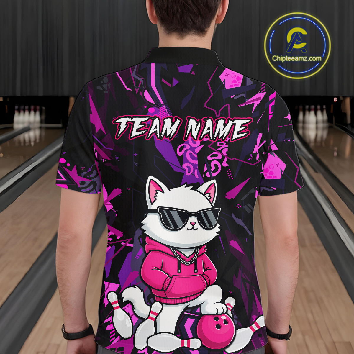 A person standing in a bowling alley wearing a short-sleeve crewneck shirt from the back. The shirt features a black base with a vibrant pink and purple graffiti-style pattern, the text 'TEAM NAME' in white with a pink outline, and a cartoon white cat wearing sunglasses and a pink hoodie with bowling pins and a bowling ball.