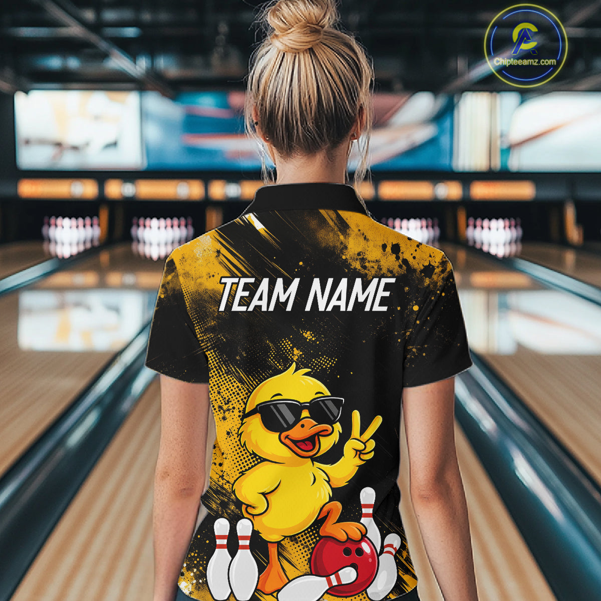 A woman viewed from behind wearing a black and yellow bowling-themed polo shirt with a cartoon duck graphic and the text 'TEAM NAME' on the back, standing in a bowling alley.