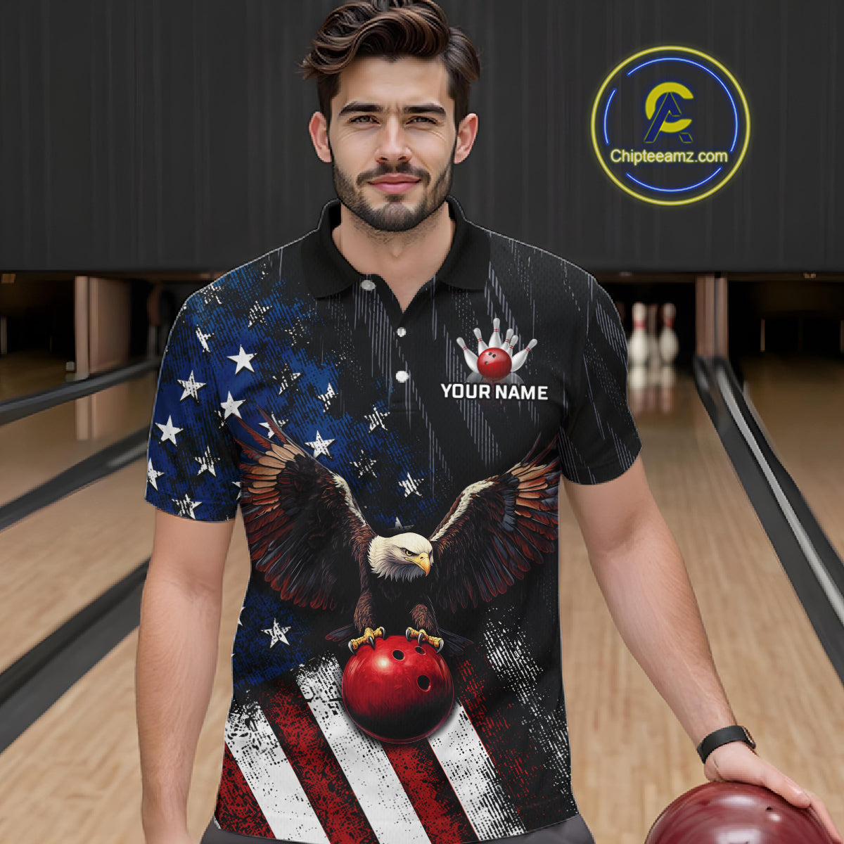 A man in a bowling alley wearing a black short-sleeve polo shirt with a patriotic American flag design, featuring a bald eagle holding a red bowling ball and white stars on the chest. He is holding a red bowling ball.