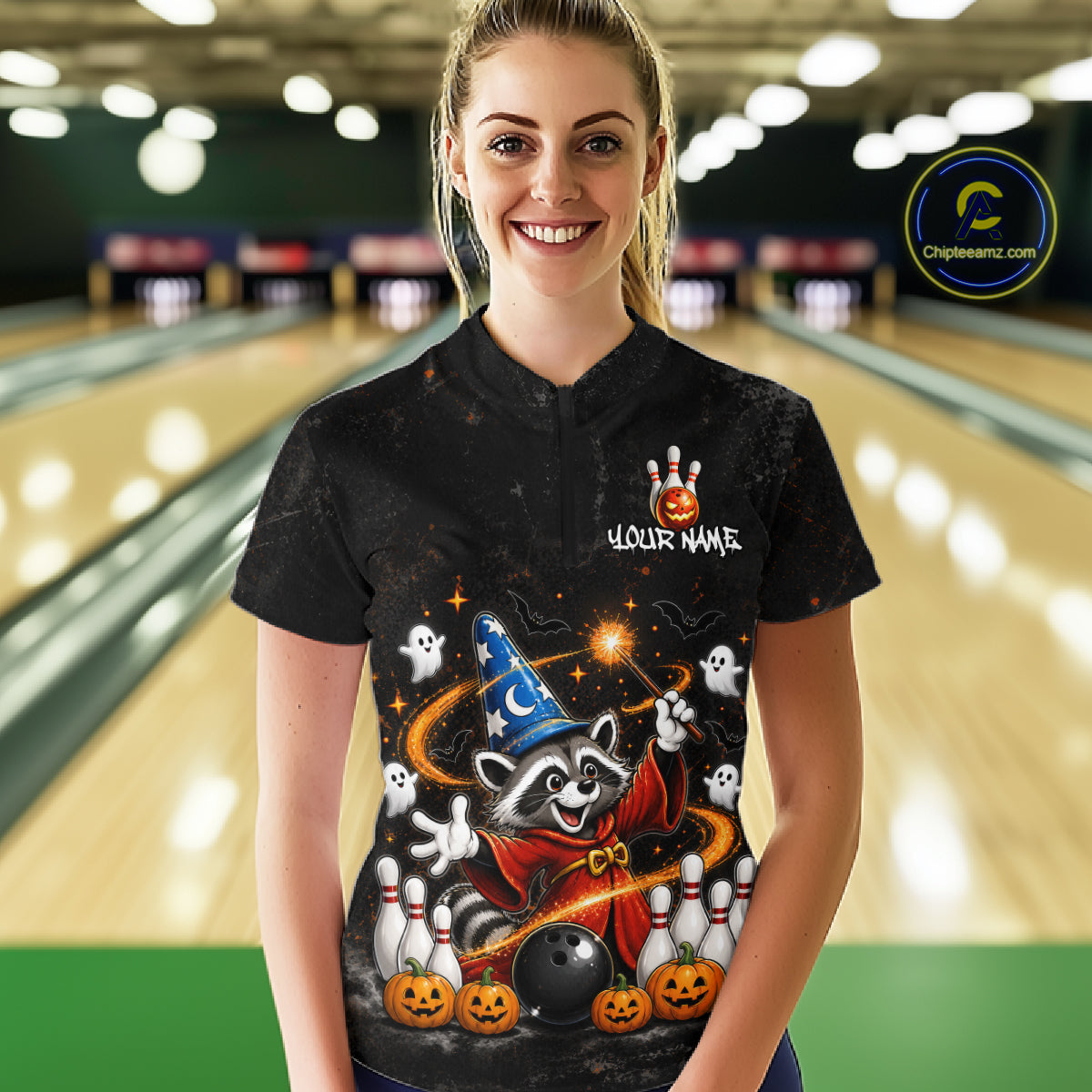 A woman stands in a bowling alley wearing a black short-sleeve polo shirt with a colorful Halloween-themed raccoon graphic on the front. The shirt features a wizard hat, ghosts, pumpkins, bowling pins, a bowling ball, and the text 'YOUR NAME' and 'Chilewich.