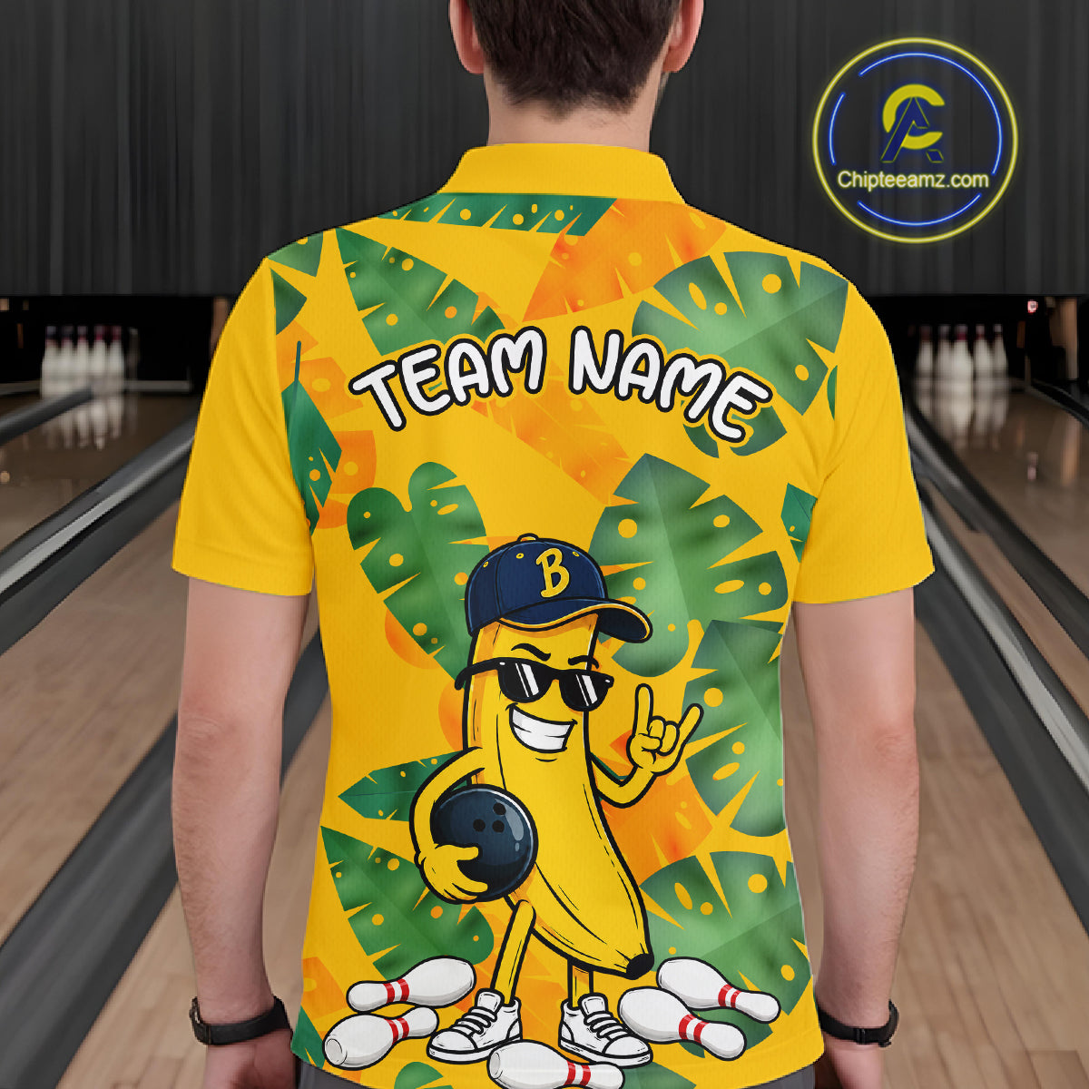 Man in a bowling alley wearing a bright yellow tropical polo shirt with green leaf patterns, custom team name text, and a smiling banana mascot holding a bowling ball.