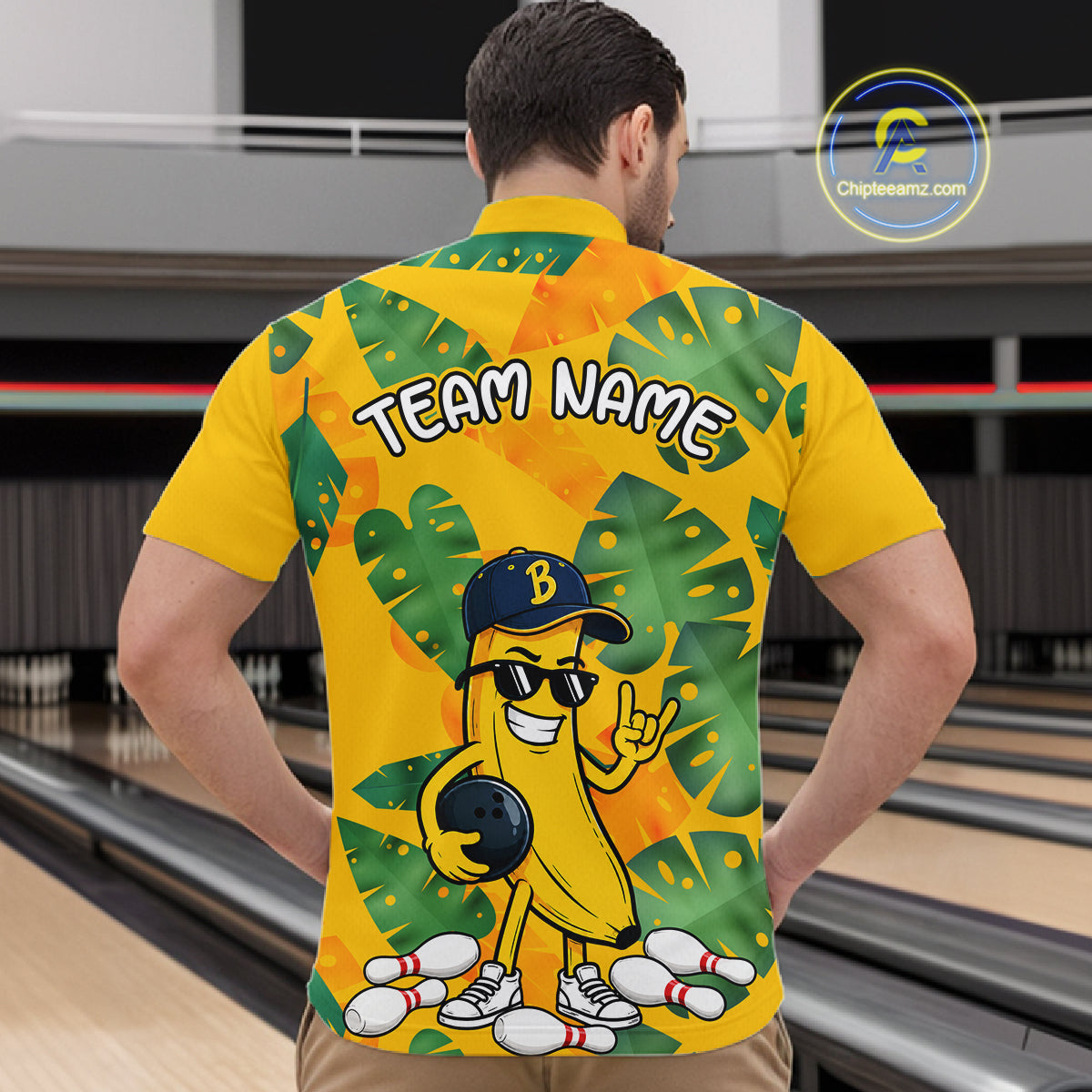 Man in a bowling alley wearing a bright yellow tropical quarter zip shirt with green leaf patterns, custom team name text, and a smiling banana mascot holding a bowling ball.