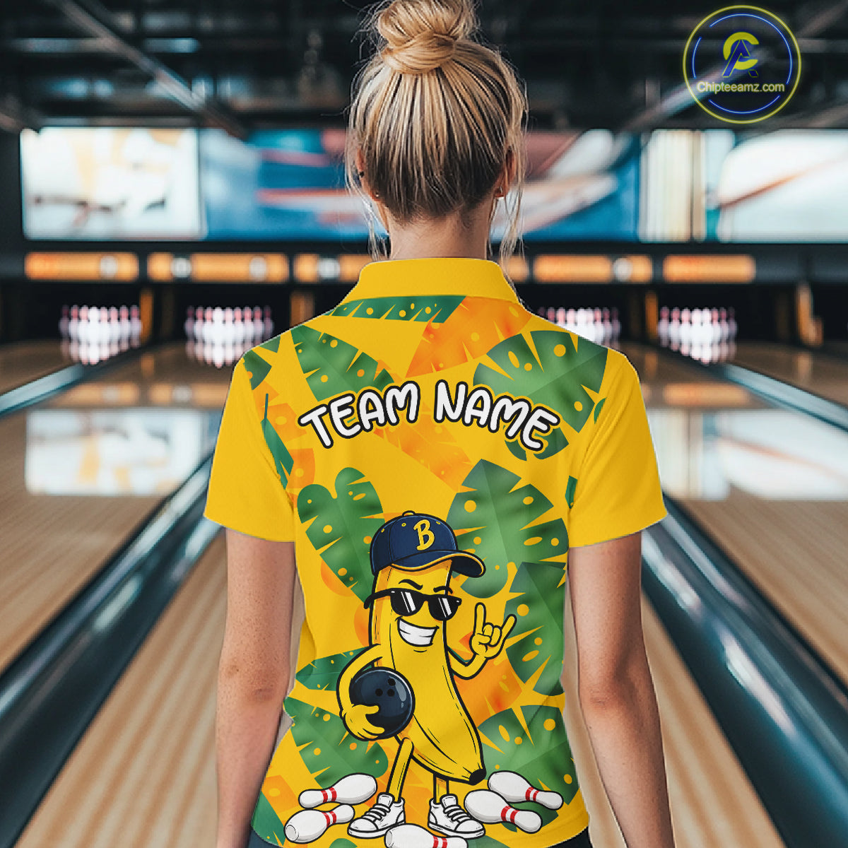 Woman in a bowling alley wearing a bright yellow tropical quarter zip shirt with green leaf patterns, custom name text, and a smiling banana mascot holding a bowling ball.
