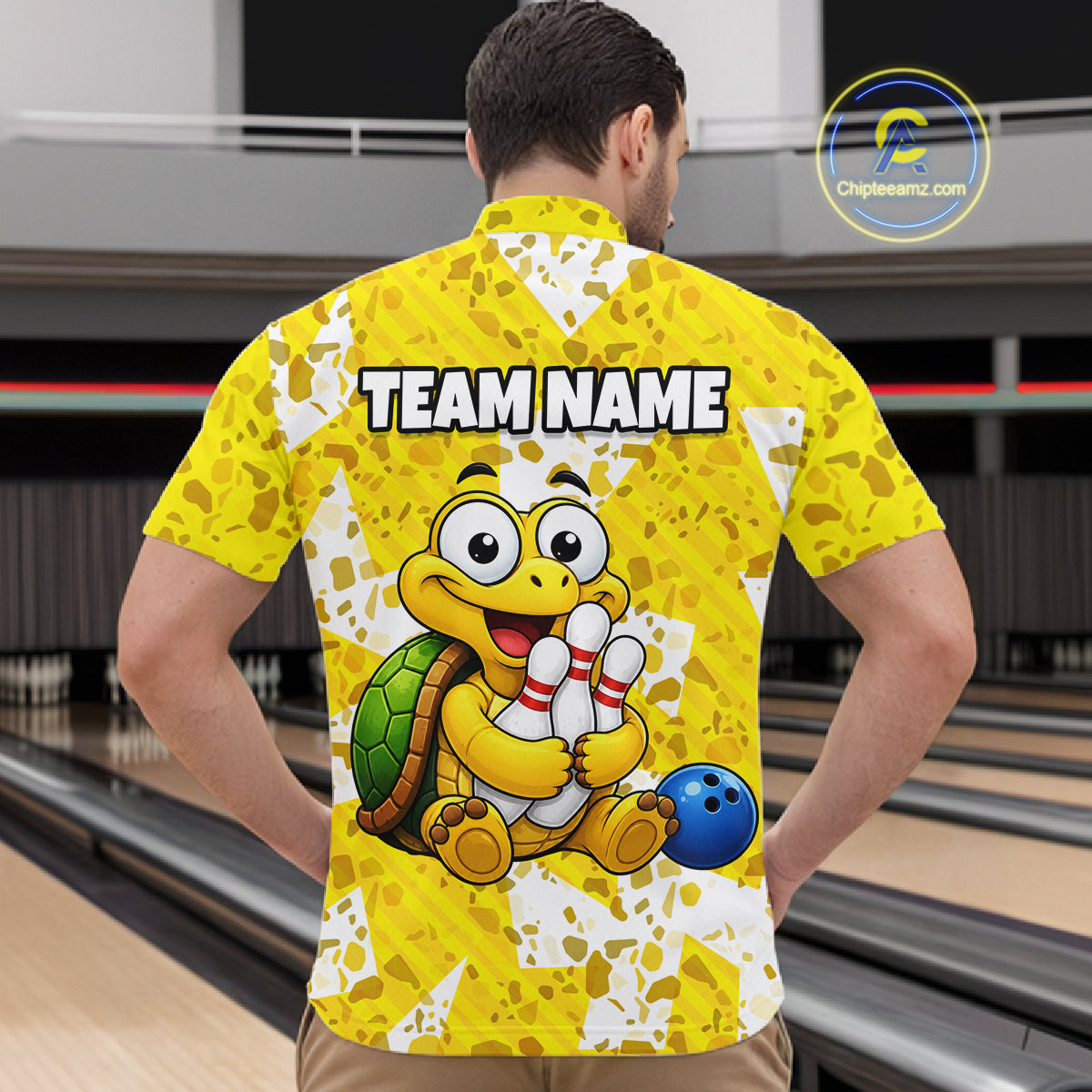 Man wearing a yellow bowling-themed shirt with a cartoon turtle holding a bowling ball in a bowling alley.