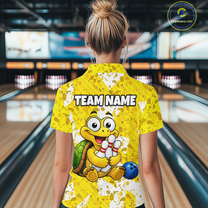 Woman wearing a yellow bowling-themed shirt with a cartoon turtle holding a bowling ball and pins in a bowling alley.