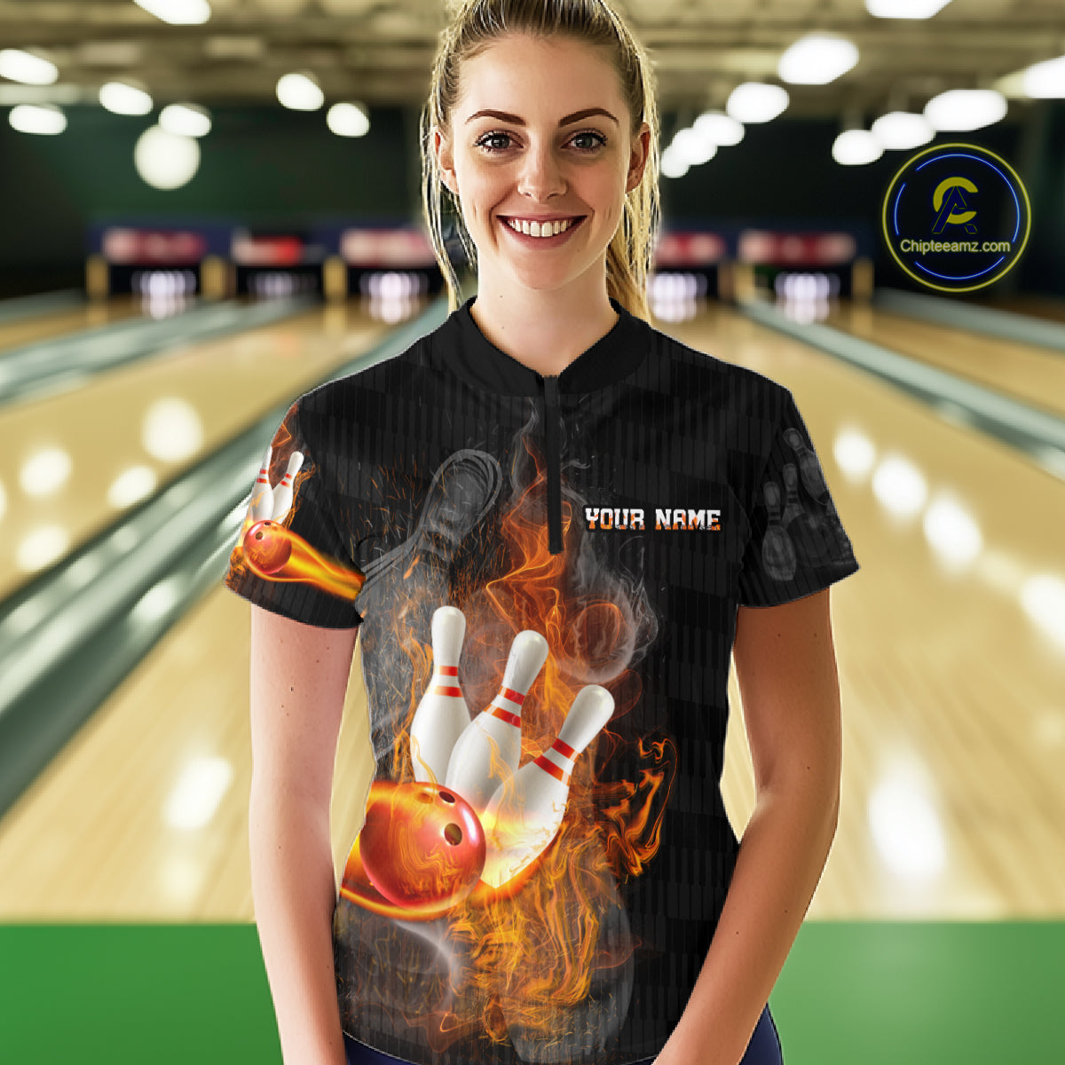 A woman stands in a bowling alley wearing a black short-sleeve custom bowling t-shirt with a graphic of a bowling ball and pins surrounded by flames, with 'YOUR NAME' printed on the chest.