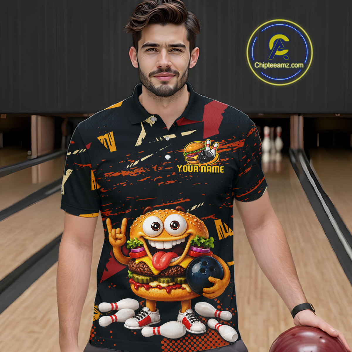 A man wearing a black bowling-themed polo shirt with a colorful cartoon burger character and bowling pins printed on the front, standing in a bowling alley holding a bowling ball. 